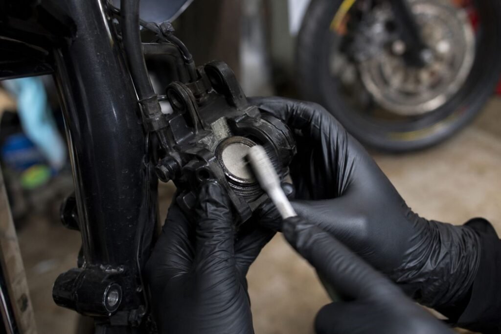 Mechanic in black gloves scrubbing a motorcycle brake caliper piston face with a nylon toothbrush without removing the caliper