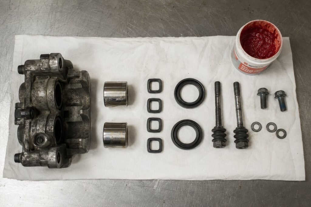 Fully disassembled motorcycle brake caliper with pistons, square-cut seals, dust seals, slide pins, and red rubber grease laid out for a step-by-step rebuild